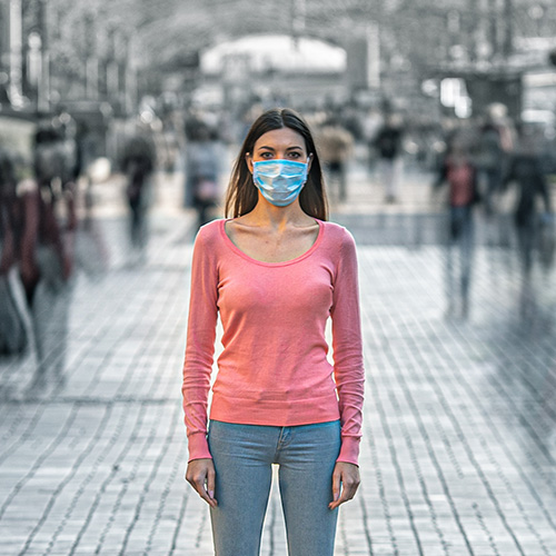 Une femme se tient debout en pleine rue. Elle a un pull rose, un jean et un masque chirugicale bleu sur la bouche et le nez. Les personnes autours sont en noirs et blanc et sont floues. Personnes n'est réellement à proximité de la femme.